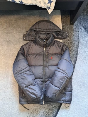 Svart pufferjacka från Polo Ralph Lauren - Säljer en svart pufferjacka från Polo Ralph Lauren med avtagbar huva och röd broderad logga på bröstet. Jackan har dragkedja framtill, två sidofickor med dragkedja och en bröstficka. Perfekt för kalla vinterdagar och riktigt skön att bära, jacka passar M-L
