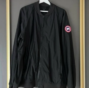 Svart bomberjacka från Canada Goose - Snygg svart bomberjacka från Canada Goose med klassisk rund krage och dragkedja framtill. Jackan har ribbade muddar och en ikonisk logotyp-patch på ärmen. Tillverkad i Portugal med meshfoder för extra komfort. Perfekt för en stilren och avslappnad look.