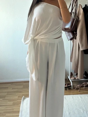 Vit off shoulder jumpsuit - Säljer ett stilrent vitt sett med one shoulder-topp och vida byxor. Toppen har en asymmetrisk skärning och knyt i midjan för en trendig look. Materialet är mjukt och luftigt ut, perfekt för en chic och avslappnad stil.