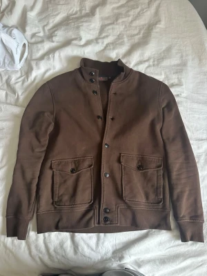 Brun kofta/cardigan från morris - Snygg brun overshirt med raka linjer och två stora fickor framtill. Jackan har hög krage, knäppning med svarta knappar och ribbade muddar vid ärmsluten. Perfekt för lager på lager och enkel att styla till olika outfits.