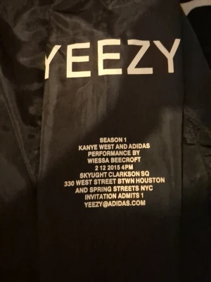 SÄLLSYNT Svart Yeezy windbreaker  - Säljer en svart hoodie från YEEZY Season 1 i samarbete med Adidas. Hoodien har vit YEEZY-logga på bröstet och detaljerad text på ryggen. Utrustad med huva och vita dragsnören samt dragkedja framtill. Materialet är glansigt och känns exklusivt.