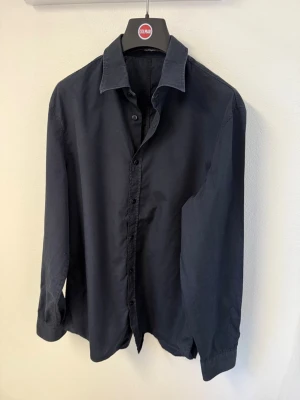 Mörkblå skjorta från J.Lindberg - Stilren mörkblå skjorta från J.Lindberg med klassisk krage och knappar framtill. Skjortan har lång ärm och är tillverkad i mjuk bomull, perfekt för en clean och enkel look. Passformen är normal och tyget är enfärgat utan mönster.