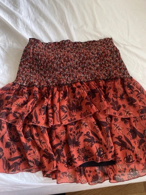 Röd volangkjol med blommönster från Zara  - Kort volangkjol i röd och svart med blommigt mönster och smockad midja. Kjolen har flera lager volanger och är gjord i ett lätt, luftigt material som passar perfekt för varmare dagar.