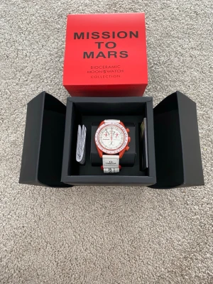 Omega x Swatch Mission to Mars - Säljer en Omega x Swatch Speedmaster MoonSwatch Mission to Mars. Klockan har en vit urtavla med röda detaljer och röd boett, samt vitt textilband med Omega- och Swatch-logga. Biokeramisk konstruktion och sportig design. Perfekt för dig som gillar unika accessoarer.