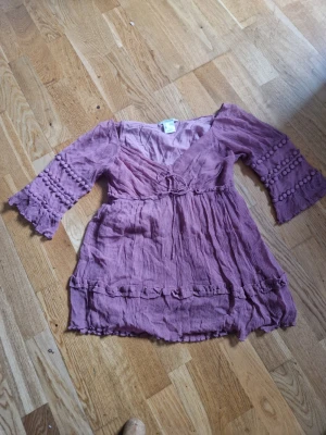 Ljuslila kortklänning med volang - Söt ljuslila kortklänning med v-ringning, knyt i midjan och volangdetaljer. Klänningen har trekvartsärm med dekorativa bollar och är gjord i ett tunt, skrynkligt bomullstyg. Perfekt för sommarens varma dagar.