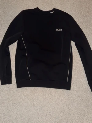 Svart sweatshirt från Hugo Boss - Stilren svart sweatshirt från Hugo Boss med rund halsringning och diskret logga broderad på bröstet. Tröjan har vita detaljer längs sidorna och ribbade muddar vid ärmslut och nederkant. Perfekt för en clean och modern look.