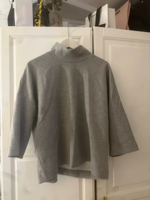 Grå polotopp från Carin Wester - Säljer en stilren grå polotopp från Carin Wester. Toppen har trekvartsärm och en hög krage som ger en clean look. Tillverkad i mjuk bomullsmix och har en loose passform som känns både chill och trendig.