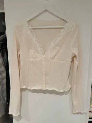 Vit spetsdetaljerad långärmad topp - Söt vit topp med lång ärm och v-ringning dekorerad med spets. Toppen har knappar framtill och är croppad med vågig kant nedtill. Aldrig använd 