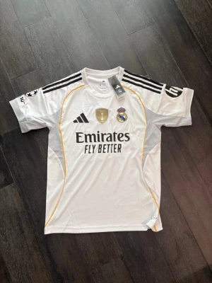 Real Madrid matchtröja Mbappé 10 - Snygg vit Real Madrid matchtröja från Adidas med svarta detaljer och guldinslag. Tryck med Mbappé och nummer 10 på ryggen. Champions League-märke på ärmen och klubbmärke på bröstet. Tillverkad i lätt och ventilerande material, perfekt för fotbollsträning eller match. Pris kan diskuteras och 