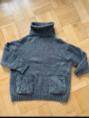 Grå polotröja med fuskpälsfickor - Mysig grå stickad polotröja med bred ribbad polokrage och två stora fuskpälsfickor framtill. Tröjan har avslappnad passform, ribbade muddar och är perfekt för kyliga dagar. Materialet känns mjukt och värmande.