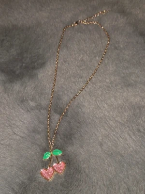 Halsband med körsbärshänge i guld - Sött halsband med ett körsbärshänge i form av två rosa hjärtan med glittereffekt och gröna blad. Kedjan är guldfärgad och hänget har en lekfull och trendig design. Perfekt accessoar för att addera lite färg och personlighet till din outfit.