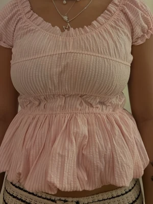 Ljusrosa ribbad blus med volang - Supersöt ljusrosa blus med ribbad struktur och volang nedtill. Toppen har korta puffärmar och smock i midjan som ger en feminin siluett. Perfekt för dig som gillar romantisk och trendig stil.