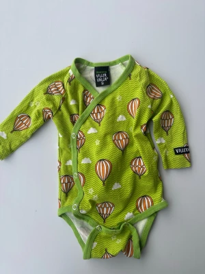 Grön body med luftballonger Viller Valla - Söt grön omlottbody från Viller Valla i ekologisk bomull. Mönstrad med orange och vita luftballonger, små moln och stjärnor på en zigzag-bakgrund. Långa ärmar och tryckknappar framtill gör den enkel att ta på och av.
