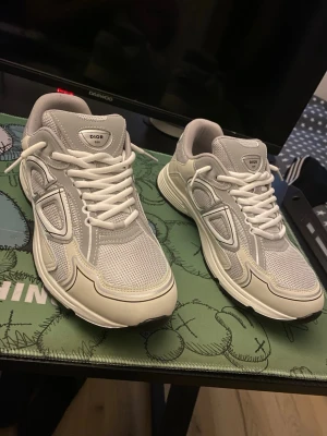 Dior B30 sneakers - Säljer ett par gråa och beiga Dior B30 sneakers med vita detaljer och meshpaneler. Skorna har chunky sula, vita skosnören och en modern, sportig design. Perfekta för dig som vill ha en lyxig och trendig look. Märket syns tydligt på plösen. Ingen BOX. Och finns en liten ljusblå fläck.