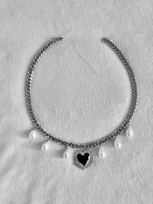 Silverfärgat halsband med pärlor och hjärta - Unikt halsband i silverfärgad metall med droppformade vita pärlor och ett svart hjärthänge med solstråleliknande detaljer runt. Kedjan är chunky och ger en cool kontrast till de eleganta pärlorna. Perfekt för dig som vill sticka ut med accessoarerna.