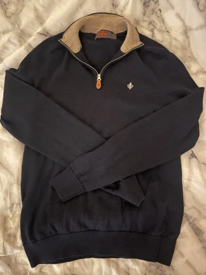 Marinblå tröja med halvzip från Morris - Snygg marinblå tröja från Morris med halvzip och beige krage. Tröjan har en liten broderad logga på bröstet och ribbade muddar vid ärmslut och nederkant. Perfekt för dig som gillar klassisk och stilren design.