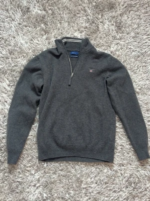 Mörkgrå halvzip tröja från Gant - Mörkgrå ull halvzip från Gant med broderad logga på bröstet. Tröjan har hög krage, långa ärmar och ribbade muddar. Perfekt för lager på lager och en stilren look. Tröjan är M men är liten i storleken passar med på någon med XS/S