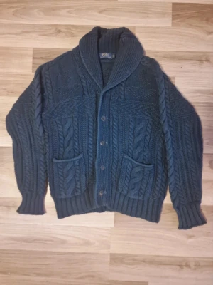 Mörkblå kabelstickad kofta Polo Ralph Lauren - Mysig mörkblå kabelstickad kofta från Polo Ralph Lauren med stora fickor framtill, grovstickad struktur och bred sjalkrage. Koftan har knäppning med fem knappar och ribbade muddar vid ärmslut och nederkant. Perfekt för lager på lager och klassisk stil. Nypris: $1098