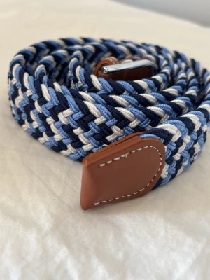 Flätat blått bälte med läderdetaljer - Snyggt flätat bälte i olika blå nyanser, vitt och mörkblått. Bältet har bruna läderdetaljer vid ändarna och ett silvrigt spänne. Perfekt för att ge din outfit en cool touch. Passar både jeans och chinos.