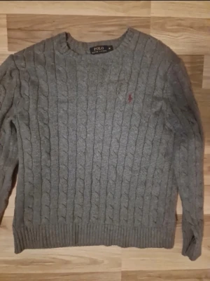 Grå kabelstickad tröja Polo Ralph Lauren - Kabelstickad grå tröja från Polo Ralph Lauren med rund halsringning och klassisk röd logga broderad på bröstet. Tröjan har ribbade muddar vid ärmslut och nederkant. Perfekt för lager på lager och en stilren look. Nypris: 2865kr