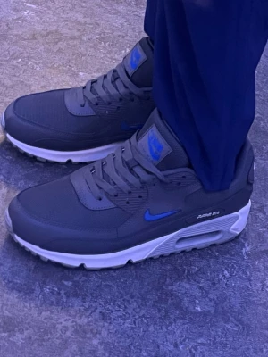 Nike Air Max 90 grå/blå sneakers - Snygga Nike Air Max 90 sneakers i mörkgrått med blå detaljer och vit sula. Ovandelen är i mesh och syntet för en sportig look. Klassisk Air Max-dämpning i sulan och ikonisk design med blå Nike-logga på sidan och plös.