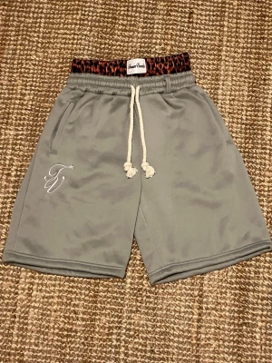 Grå shorts med dubbel midja och tjocka snören - Snygga grå shorts med dubbel midja och vit snörning i midjan och leopardmönstrad resår. Shortsen har två sidofickor och broderad logga på ena benets framsida och baksidan. Tillverkade i mjukt syntetmaterial, perfekta för sommarens alla äventyr. Shortsen har en bra passform och är perfekt för någon som är intresserad av fashion