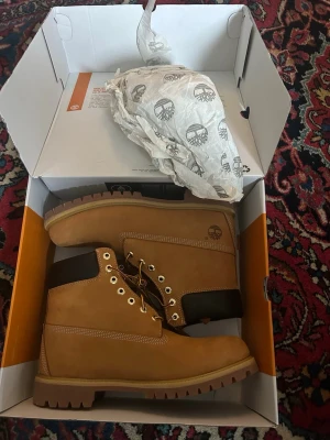 Timberland klassiska kängor i nubuck - Säljer ett par klassiska Timberland kängor i ljusbrun nubuck med robust sula och mörkbrunt skaft. Kängorna har guldfärgade öljetter, grova snören och den ikoniska Timberland-loggan på sidan. Perfekta för dig som vill ha en tidlös och street-inspirerad look.