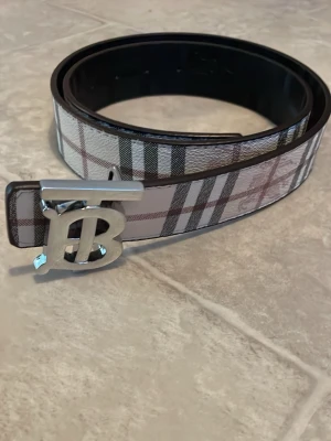 Rutig Burberry skärp med silverdetalj - Snyggt rutig Burberry skärp med klassiskt mönster i vitt, svart och brunt. Kan användas på båda håll. Skärpet har ett stort, blankt silverspänne med Burberrys ikoniska B-logga. Perfekt accessoar för att lyfta vilken outfit som helst.