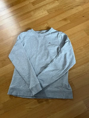 Ljusgrå sweatshirt från Gant - Ljusgrå sweatshirt från Gant med rund halsringning och diskret logga broderad på bröstet. Tröjan har långa ärmar, ribbade muddar och är tillverkad i mjukt bomullsmaterial. Perfekt för en avslappnad och stilren look.