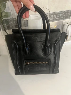 Svart Celine Nano Luggage handväska - Säljer en svart Celine Nano Luggage handväska. Väskan har två topphandtag, avtagbar axelrem och en liten ficka med dragkedja framtill. Klassisk och stilren design med diskret Celine Paris-logga. Skick: relativt bra skick men en del sprucket läder på insidan av väskan. Lite guld på Celine loggan har nötts av. Väskan skickas med en Celine dustbag. Köpt begagnad på Vinted för några år sedan så tyvärr har jag ej kvitto eller liknande. 