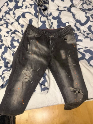 Svart/Grå dsquared2 jeans med färgstänk - Säljer ett par svarta jeans från Dsquared2 med slitna detaljer och coola färgstänk i orange och vitt. Jeansen har en relaxed fit. Perfekta för dig som gillar en edgy och trendig stil.