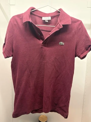 Vinröd pikétröja från Lacoste - Vinröd pikétröja från Lacoste med klassisk krage, korta ärmar och knappar vid halsen. Ikoniska gröna krokodilloggan på bröstet. Tillverkad i mjuk bomull för en skön och avslappnad stil.