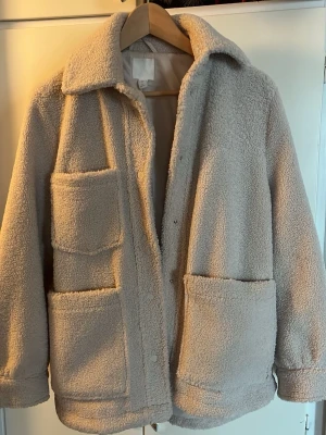 Beige teddyjacka från H&M XS - Mysig beige teddyjacka från H&M i storlek XS. Jackan har två stora framfickor, klassisk krage och stängs med tryckknappar. Tillverkad i mjukt teddy-material som ger en avslappnad och trendig look. Perfekt för kyliga dagar och enkel att matcha med olika outfits.