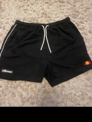 Svarta shorts från Ellesse med snörning - Svarta shorts från Ellesse med vit snörning i midjan och meshfoder. Klassisk Ellesse-logga på vänster ben och en patch med märkesnamnet på höger ben. Perfekta för sommaren och sportiga aktiviteter.