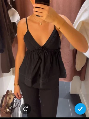 Svart linnetopp med knyt och spets - Snygg svart linnetopp med tunna axelband, v-ringning och spetsdetaljer vid bysten. Toppen har knyt framtill och en lös, flowy passform som ger en avslappnad look. Perfekt för varma dagar eller att styla lager på lager.