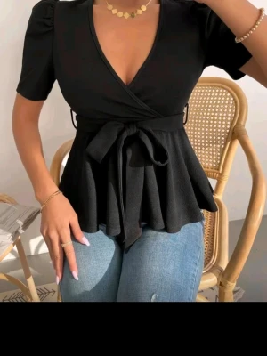 Svart topp med knytband - Stilren svart topp med v-ringning och korta puffärmar. Toppen har ett brett knytband i midjan som markerar figuren och ger en snygg siluett, perfekt till jeans eller kjol.