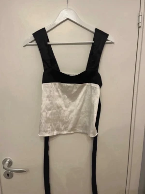 Svartvit korsettopp med breda band - Snygg svartvit korsettopp med breda axelband och långa knytband i sidan. Toppen har en vit kropp i glansigt material och svarta detaljer upptill och på banden. Perfekt för att lyfta vilken outfit som helst.