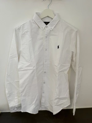 Vit skjorta från Ralph Lauren M - Klassisk vit skjorta från Ralph Lauren med button-down krage och broderad logga på bröstet. Skjortan är långärmad och har knappar framtill. Tillverkad i mjuk bomull för en clean och tidlös look.