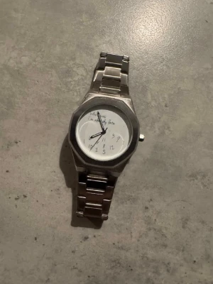 Silverfärgad klocka med länkarmband - Stilren klocka i silver med metallänk och vit urtavla. Urtavlan har handskrivna siffror och text, vilket ger en unik och personlig touch. Klassisk design som passar till många olika stilar.