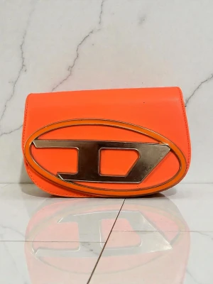 Orange kuvertväska från D - Snygg orange kuvertväska från Dx i slätt läder. Väskan har en unik oval gulddetalj med D-logga på framsidan som verkligen sticker ut. Perfekt för dig som vill ha en färgklick till outfiten och gillar statement-accessoarer.