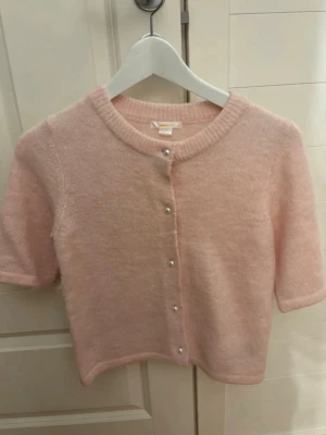 Ljusrosa kofta från Gina Tricot - Supersöt ljusrosa kofta från Gina Tricot med korta ärmar och pärlknappar framtill. Koftan är stickad i ett mjukt material och har en rund halsringning. Perfekt att slänga över en topp för en gullig look.