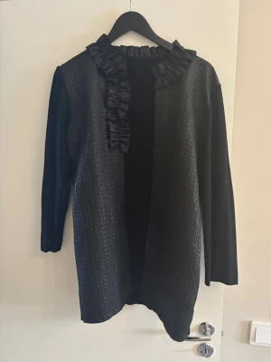 Svart kofta med volangkant - Stilren svart kofta med öppen front och dekorativ volangkant längs halsringningen. Koftan har lång ärm och är tillverkad i ett mjukt, lätt glansigt material framtill och slät baksida. Perfekt för att lyfta en enkel outfit.