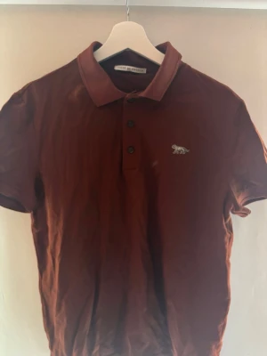 Brun pikétröja från Tiger of Sweden - Snygg brun pikétröja från Tiger of Sweden med klassisk krage och knappar framtill. Tröjan har korta ärmar och en liten broderad logga på bröstet. Perfekt till jeans eller chinos för en clean look.