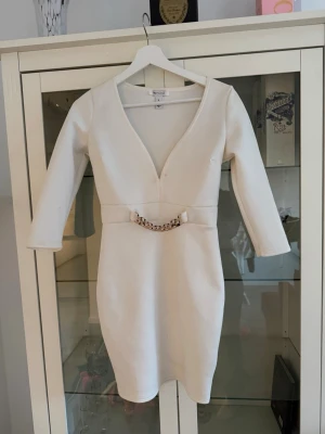 Vit kortklänning med kedjedetalj - Säljer en stilren vit kortklänning från NLY One med v-ringning, trekvartsärm och en snygg guldfärgad kedjedetalj i midjan. Klänningen har figurnära passform och är tillverkad i ett stretchigt material som ger en smickrande siluett.