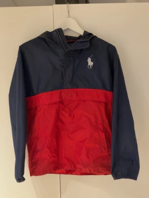  Ralph Lauren - Snygg vindjacka i marinblått och rött från Polo Ralph Lauren. Jackan har huva, dragkedja vid halsen och stor logga med flaggor på ryggen. Klassisk Polo-häst på bröstet. Tillverkad i lätt, vindtätt material som passar perfekt till streetwear.