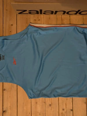 Blå ärmlös linnetopp från Kalenji - Snygg blå ärmlös linnetopp från Kalenji med vit och röd rand längs sidan. Tillverkad i lätt och ventilerande polyester, perfekt för aktiva dagar. Diskret logga på bröstet i rött. Enkel och sportig design som passar till träning.