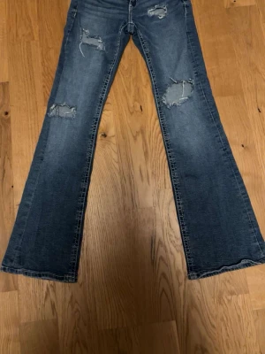 Blå slitna bootcut jeans - Säljer ett par blå bootcut jeans med slitningar och hål på framsidan. Jeansen har en klassisk femficksmodell och är tillverkade i denim. Perfekta för dig som gillar en avslappnad och trendig stil.Innerbenslängd 74 midja 33, storlek 158💙