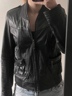 Svart skinnjacka med dragkedjor - Snygg svart skinnjacka med ribbade muddar i midjan och vid kragen. Jackan har två framfickor med dragkedjor och markerade axelpartier. Stängs med dragkedja framtill och har en klassisk, tidlös look.