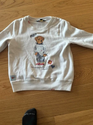 Vit sweatshirt Polo Bear Ralph Lauren - Vit sweatshirt från Ralph Lauren med det ikoniska Polo Bear-trycket framtill. Tröjan har rund hals, ribbade muddar och detaljer som broderade hjärtan och stjärna. Tillverkad i mjukt bomullsmaterial för en skön och avslappnad stil. Tröjan är för barn storlek 12 år