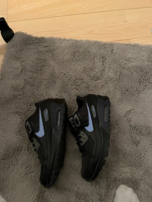 Nike Air Max 90 svart/blå sneakers - Säljer ett par Nike Air Max 90 sneakers i ny skick i färgen svart med ljusblå swoosh och detaljer. Skorna har klassisk chunky sula med synlig Air-enhet och ovandel i en mix av läder och mesh. Perfekta för dig som gillar streetwear och sportig stil.